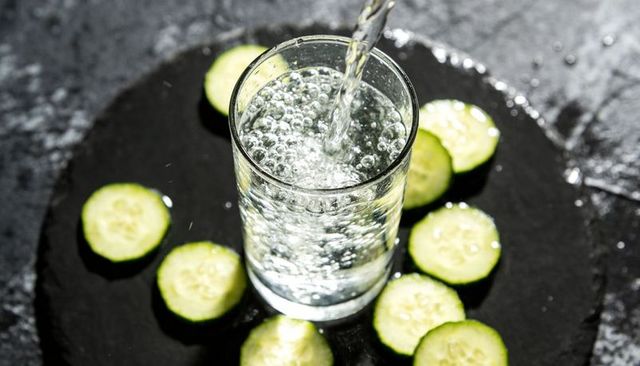 Pouring sparkling water into glass with cucumber slices on dark slate surface for healthy drink