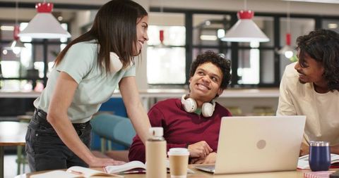 Sunlit university students collaborating around laptop with headphones in modern campus