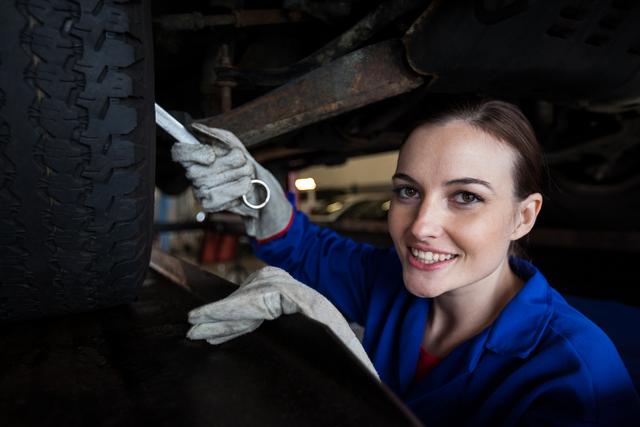 Woman mechanic smiling and using a wrench to service a car in a repair garage. Useful for content related to women in automotive industry, professional mechanics, car maintenance services, and gender diversity in technical fields.