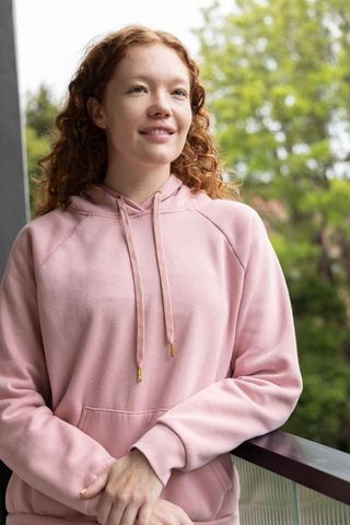 Red-haired woman standing balcony wearing pink hoodie, leaning on railing, feeling serene
