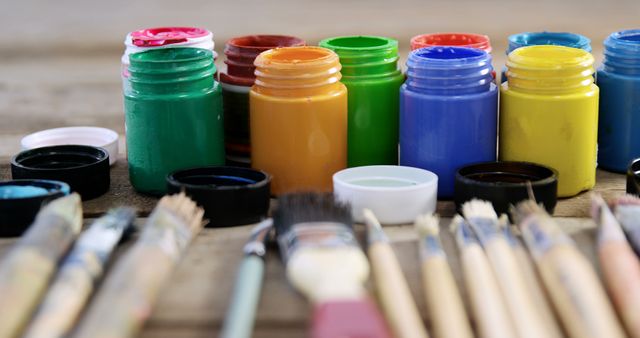 Vibrant acrylic paint jars in a variety of colors are arranged with paintbrushes on a wooden table. This image is perfect for promoting art supplies, creative projects, or hobby-related content. Use it in online shops, blogs about painting techniques, or educational materials.