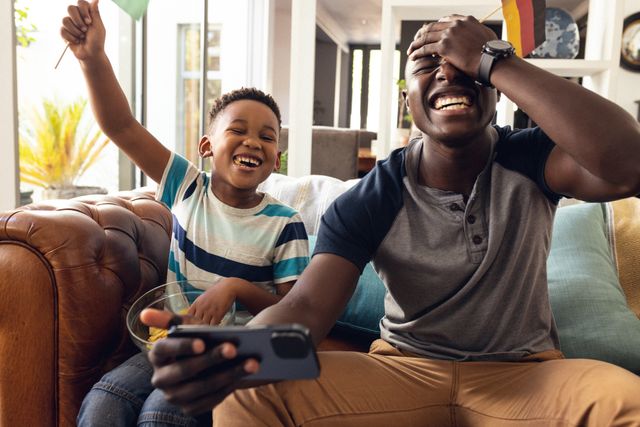 Happy african american father and son having fun playing video game together on couch in living room. Enjoying quality family time together at home with technology.