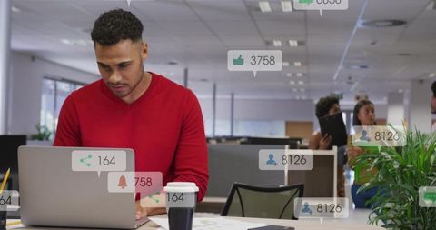 Man in red sweater typing on laptop with social engagement badges in modern open office