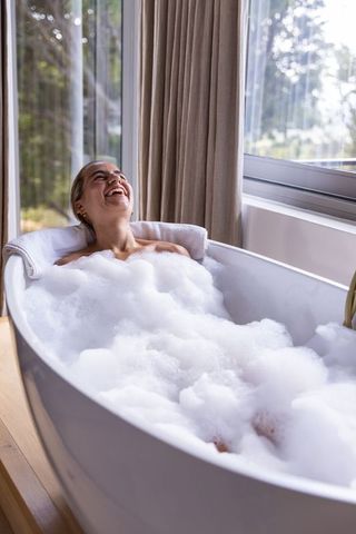Woman relaxing in freestanding bubble bath by window laughing in natural light retreat