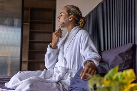 Woman in white bathrobe using jade facial roller while relaxing on bed in minimalist bedroom