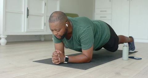 African american man performing forearm plank at home wearing earbuds and smartwatch