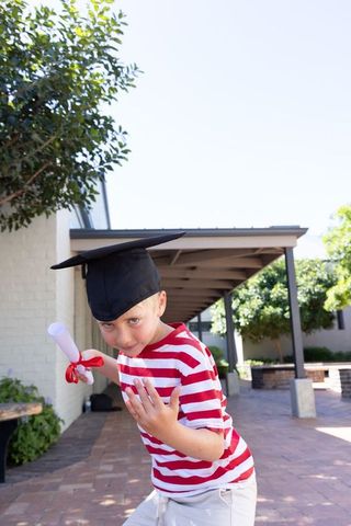Playful young graduate wearing mortarboard and red striped shirt holding diploma outdoors