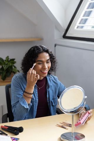 Non-binary person applying makeup at sunlit loft desk using mirror, brushes, palette