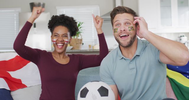 Mixed-race couple sitting in living room, cheering and celebrating while watching football match on television. Man and woman have painted flags on faces, holding football surrounded by sports memorabilia and flags. Perfect for ads, social media posts, or blog articles about sports enthusiasts, unity in sports, or home entertainment.