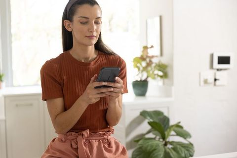 Mid-adult woman sitting on kitchen counter using smartphone in bright minimalist home