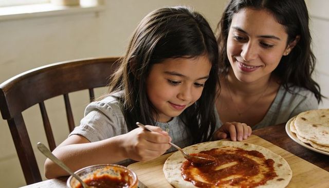 Mother guiding daughter spreading tomato sauce on pita during home cooking and meal prep