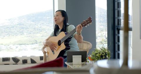 Asian woman playing acoustic guitar in cozy sunlit living room, musician practicing