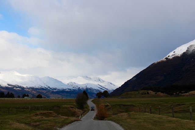 A quiet road stretches into the distance between snowy mountain ranges and green meadows under a partly cloudy sky. Ideal for use in promoting travel destinations, adventure tourism, and picturesque landscapes. Can be used in nature magazines, travel blogs, and countryside retreats advertisements.