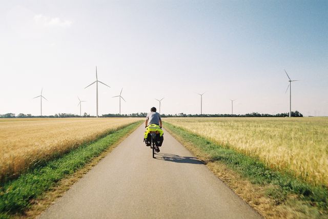 Cyclist riding bike along paved rural road surrounded by open fields with multiple wind turbines on horizon. Ideal for use in content related to sustainable travel, renewable energy, green living, cycling tourism, and outdoor adventures.