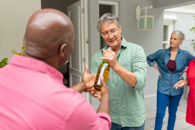 African american senior man giving wine bottle to caucasian male friend at entrance of house. unaltered, lifestyle, friendship, house party, arrival and leisure.