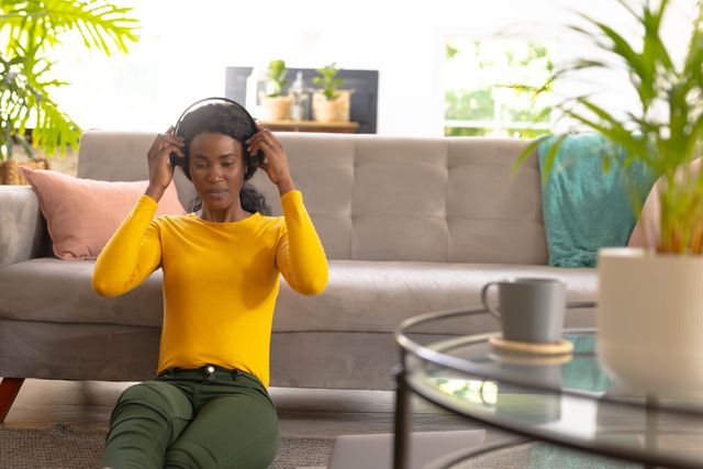 Young African American woman enjoying music with headphones while sitting on the floor against a sofa in a modern living room. Ideal for use in lifestyle blogs, technology advertisements, home decor magazines, and relaxation-themed content.