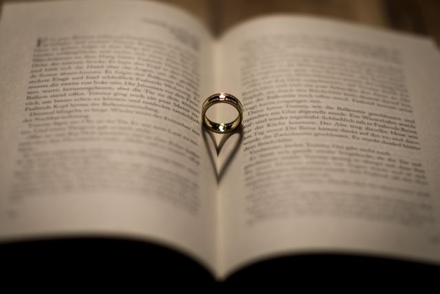 Golden ring casts heart-shaped shadow on pages of open book, symbolizes love and romance. Perfect for illustrating themes of relationship, wedding, literature and Eros. Effective for use in greeting cards, wedding invitations, romantic decorations, engagement announcements and Valentine's Day promotions.