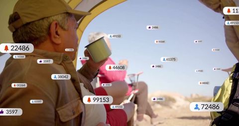 Senior couple camping on beach dunes checking social notifications while sipping coffee