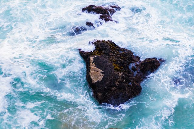 Aerial view of waves crashing against dark rocks surrounded by vibrant turquoise water. Ideal for marine life articles, oceanic conservation campaigns, travel brochures, or background enhancements for aquatic themes. Visualizes power and beauty of natural seascapes.