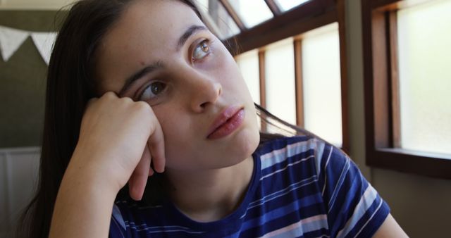 Teenage girl with long hair resting her head on hand, daydreaming while looking away. She is wearing a casual striped shirt, seated indoors near a window with natural light illuminating her face. Suitable for themes of adolescence, pondering, introspection, or personal growth.