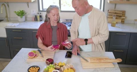 Asian senior couple preparing colorful fruit platter and baguette on marble kitchen island