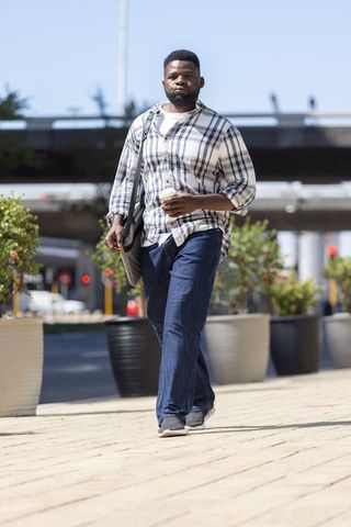 African man walking with coffee and shoulder bag along city sidewalk under overpass