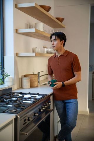 Asian man holding green mug gazing out window in sunlit kitchen with stainless range