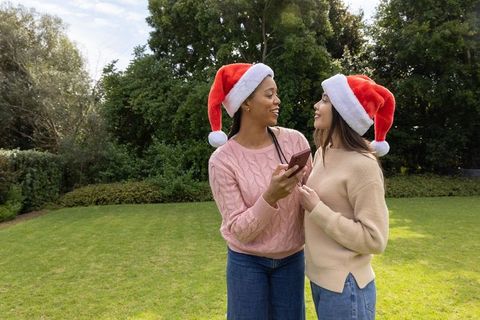 Diverse women celebrating holidays outdoors wearing santa hats and using smartphone