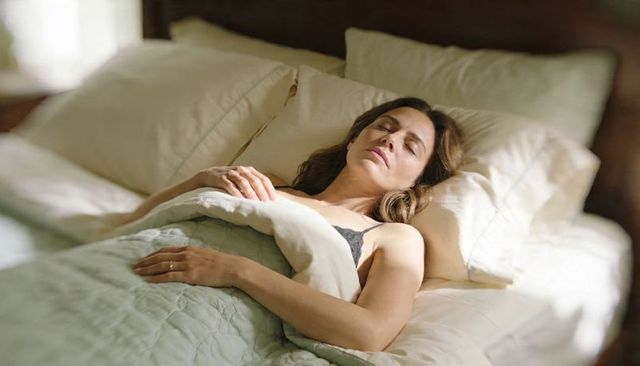 Woman resting in sunlit bed with layered bedding and stacked pillows, peaceful morning