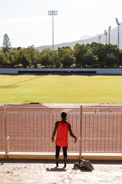 Rear view of fit, biracial disabled male athlete at an outdoor sports stadium, preparing for workout in the stands wearing headphones and running blades. Disability athletics sport training.