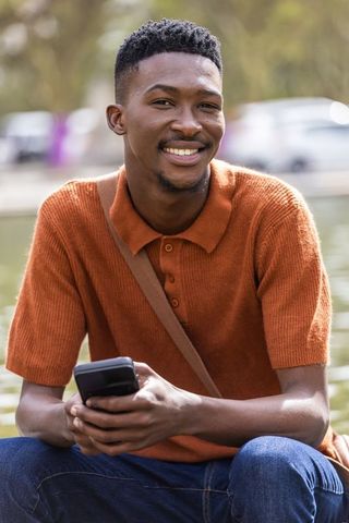 Smiling african man by lake holding smartphone, rust polo, crossbody strap, relaxed lifestyle