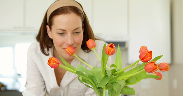 Woman happily admiring a vibrant bouquet of orange tulips on a kitchen countertop. The bright setting and her serene expression create an atmosphere of tranquility and home comfort. Perfect for advertising home decor, lifestyle blogs, gardening magazines, and springtime promotions.