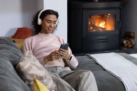 Non-binary person relaxing on sofa listening with wireless headphones and using smartphone