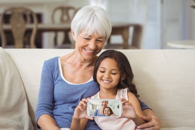Grandmother and granddaughter sitting on couch in living room, smiling and taking selfie with mobile phone. Perfect for use in family-oriented advertisements, technology promotions, or articles about intergenerational relationships and bonding.