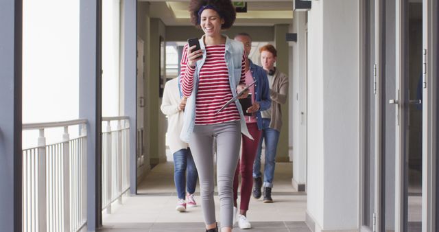 Young student strolling through a building corridor, happily looking at her smartphone and smiling. Ideal for educational websites, college brochures, student life blogs, and promotional materials for academic institutions.