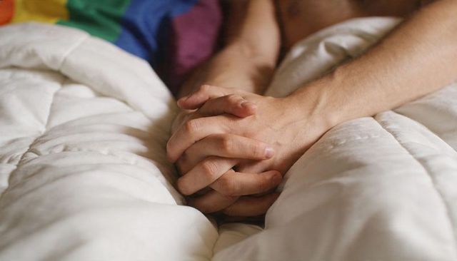 Clasped hands couple resting in bed under white duvet with rainbow throw intimate morning