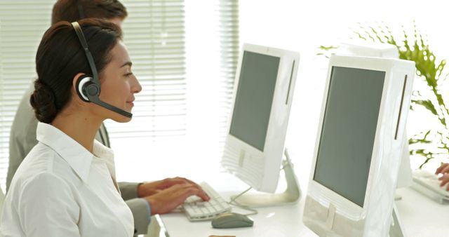 An Asian woman wearing a headset is working at a computer in an office environment, with copy space. Her profession is in customer service or a call center, indicated by the headset and computer setup.