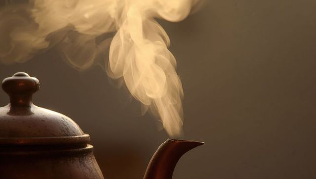 Bronze teapot spout releasing wispy steam backlit amber macro showing patina and vapor