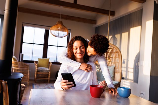 Happy, diverse lesbian couple using smartphone and embracing at table in kitchen - Download Free Stock Photos Pikwizard.com