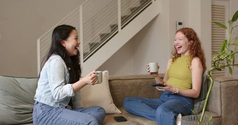 Friends laughing and sharing coffee while scrolling smartphone on cozy modern sofa