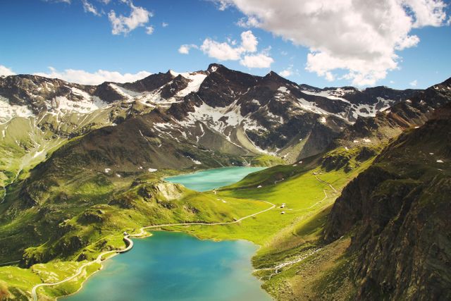 Scenic view of alpine mountain landscape featuring lush green valleys, emerald lakes, and snow-capped peaks under a blue sky with clouds. Ideal for promoting travel, adventure activities, nature exploration, and outdoor recreation. Suitable for use on tourism websites, nature blogs, outdoor gear advertisements, and posters about hiking and relaxation in natural settings.