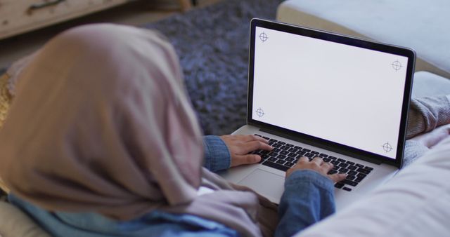 Over shoulder image of biracial woman in hijab at home working on laptop, copy space on screen. Remote working, communication, inclusivity and domestic life.
