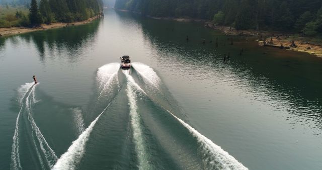 This image shows an aerial view of a wakeboarder being pulled by a speedboat in a calm lake surrounded by forested hills. The scene creates a sense of adventure and fun, making it ideal for promoting outdoor activities, summer vacations, or advertising water sports equipment. Perfect for travel brochures, sports websites, and adventure blogs.
