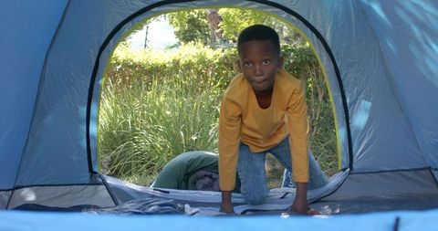Young boy crawling through blue tent doorway during backyard camping under sunlight