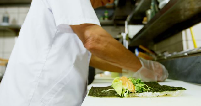 Chef wearing gloves in a restaurant kitchen preparing a fresh sushi roll with nori and various ingredients. Useful for content related to culinary arts, Japanese cuisine, restaurant businesses, food preparation techniques, and professional chef skills.