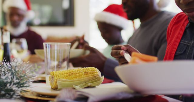 Close-up of a family enjoying a Christmas dinner with a focus on corn on the cob on the table. Family members in festive hats gathered around table, celebrating holiday together. Ideal for illustrating holiday gatherings, festive meals, family moments.