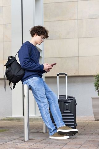 Teen traveler leaning on bollard checking smartphone with carry-on suitcase and backpack