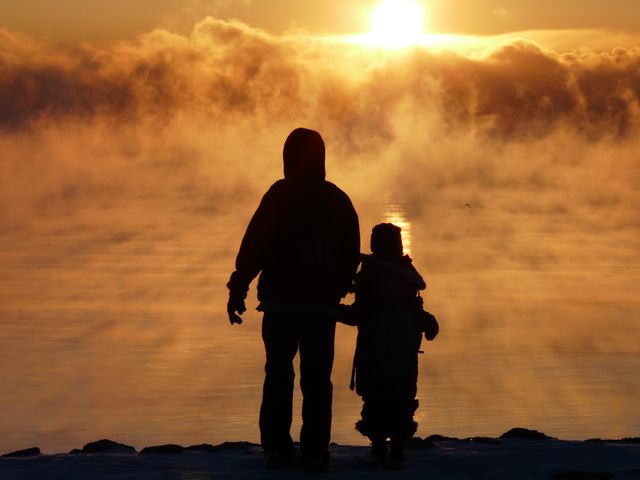 Silhouetted parent and child standing by misty lake during sunset, creating a serene and tranquil scene with mist rising. This image can be used for themes of family bonding, nature, peaceful moments, or outdoor activities. It is ideal for use in family-oriented campaigns, mental wellness, or lifestyle promotional materials.