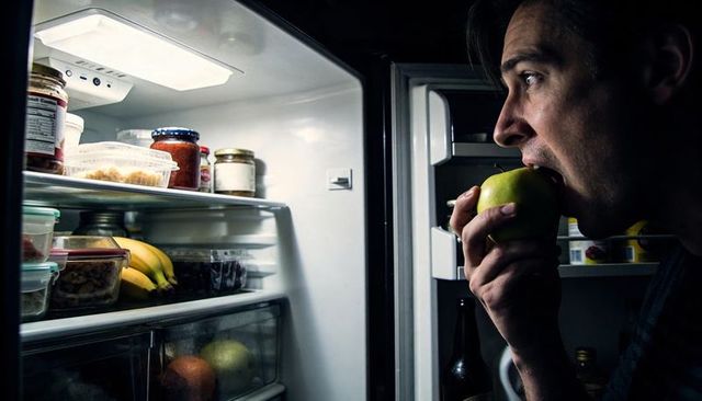 Man biting green apple while looking into open fridge at night, midnight snacking
