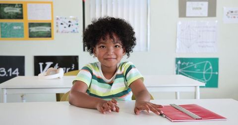 African American child sitting at classroom desk smiling with red notebook and ruler